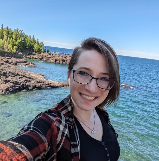 Photograph of Hilary Severyn, a white woman with short brown hair and glasses, in front of a rocky lakeshore.