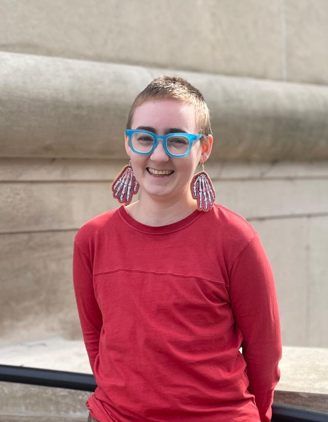 A white person with buzzed dark blonde hair, wearing chunky electric blue glasses, large embroidered skeleton hand earrings, and a long sleeve red shirt smiling with teeth