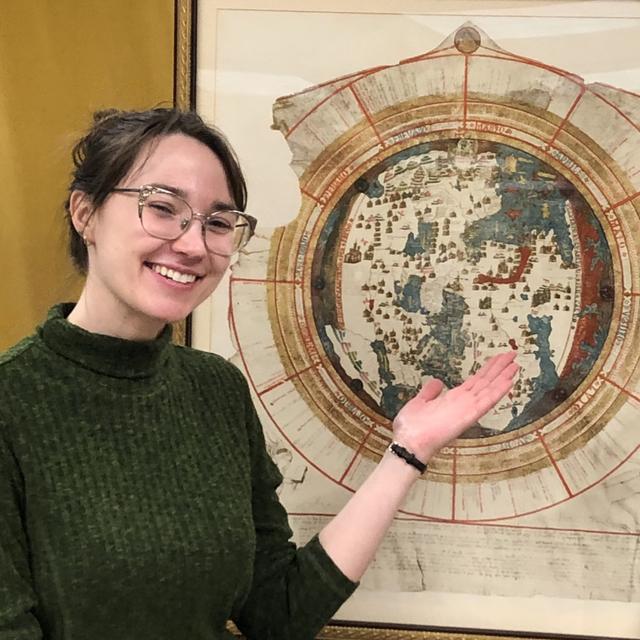 A white woman in a green turtleneck gestures behind her towards a circular map of the world from 1542.