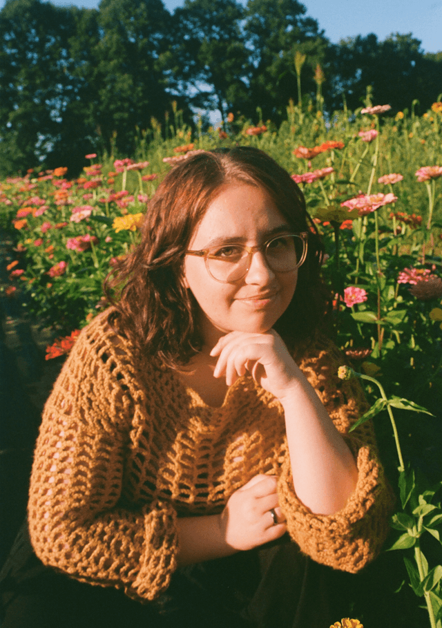 Young adult woman kneeling in a field of flowers during daytime and making eye contact with the viewer. She is wearing a yellow crocheted shirt and resting her head on her hand.