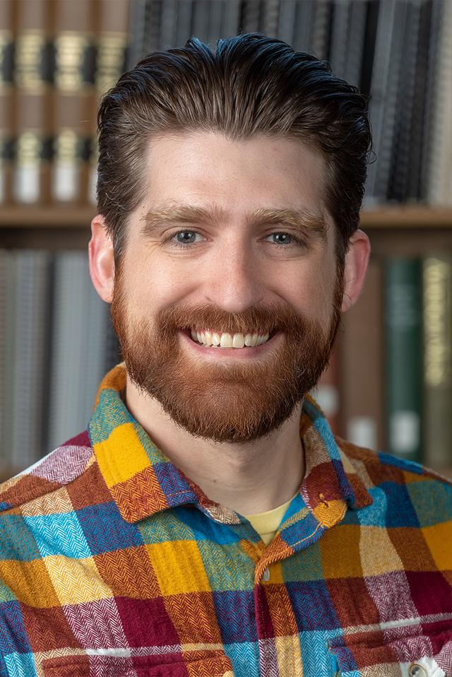 Photograph of Patrick Goussy, a white man with short brown hair and beard, wearing a plaid shirt and smiling.