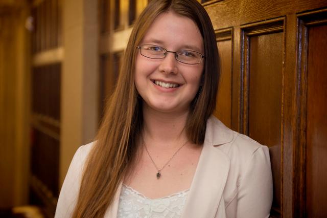 A young, white woman with chest length hair and wearing glasses smiles while leaning against a wood paneled wall. She is wearing a suit jacket, a camisole, and a small pinecone necklace. 