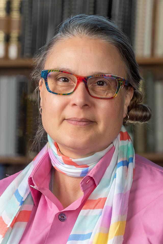 Photo of Marilène &quot;Mars&quot; De Ritis - a woman with greying hair and multi-colored eyeglass frames wearing a magenta shirt and light-weight scarf in front of Reference Reading Room bookshelf