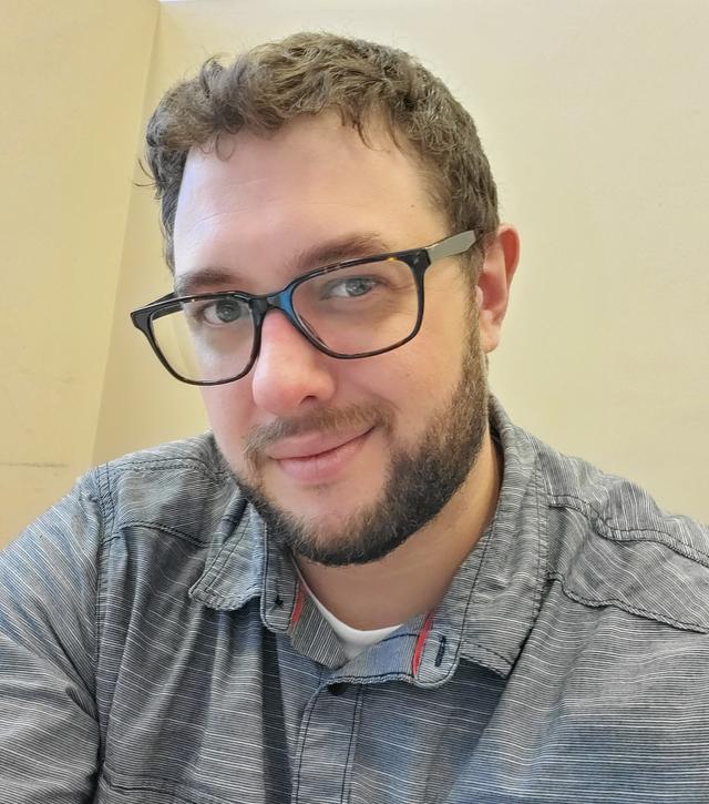 Photograph of Erik LaCoss, a white male with short brown hair, brown glasses in a grey button down shirt. 